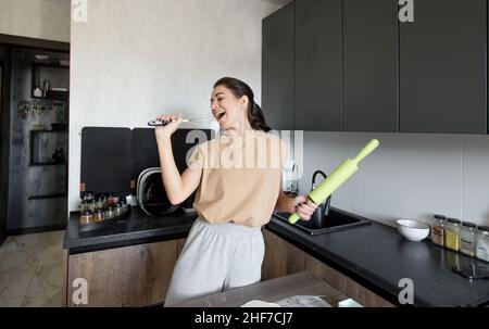 una ragazza che si diverte in una cucina moderna a casa, una ragazza felice che tiene una frusta come microfono, Foto Stock