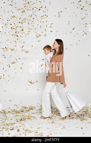 tutta la lunghezza della madre felice che tiene i sacchetti dello shopping e il figlio del toddler nelle braccia vicino ai confetti sul grigio Foto Stock