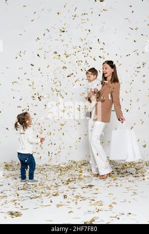 felice madre che tiene borse shopping e ragazzo toddler nelle braccia vicino alla ragazza con sindrome di down e confetti su grigio Foto Stock