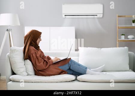 Donna sdraiata sul divano utilizzando Laptop godendo il raffreddamento del condizionatore d'aria di casa Foto Stock