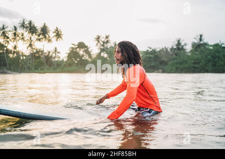 Teen uomo nero dai capelli lunghi che galleggia su una lunga tavola da surf, in attesa di un'onda pronta per il surf con palme boschetto litted al tramonto raggi. Sport acquatici estremi a Foto Stock