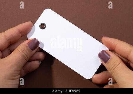 Le mani femminili tengono cartoncino bianco rettangolare vuoto etichetta con piccolo foro nella parte superiore per vestiti al centro di sfondo marrone. Tag mock up. Copia Foto Stock