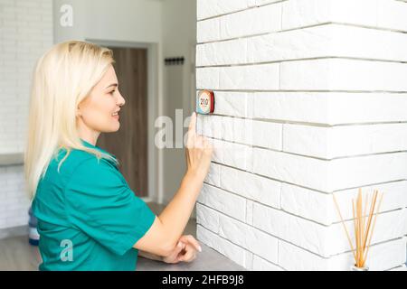 donna che regola la temperatura di riscaldamento con un moderno termostato wireless installato sulla parete bianca a casa. Concetto intelligente di regolazione del riscaldamento domestico Foto Stock