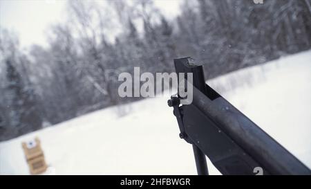 Un uomo che punta una pistola a pellet verso un bersaglio, praticando il suo obiettivo in inverno. Clip. Lo sparatutto in camouflage mira a un bersaglio con un fucile con un'op Foto Stock