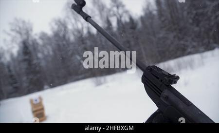 Un uomo che punta una pistola a pellet verso un bersaglio, praticando il suo obiettivo in inverno. Clip. Lo sparatutto in camouflage mira a un bersaglio con un fucile con un'op Foto Stock