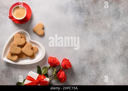 Caffè di San Valentino e biscotti a forma di cuore in piatto, bouquet di rose rosse e regalo d'amore. Vista dall'alto. Spazio di copia. Foto Stock