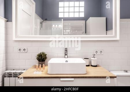 Vista frontale del guardaroba con lavello quadrato in porcellana sul piano di lavoro in legno e specchio incorniciato bianco sulla parete di piastrelle bianche Foto Stock