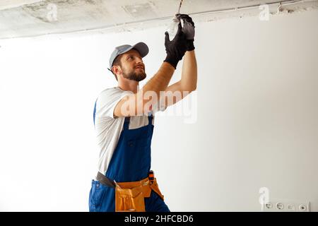 Elettricista concentrato in uniforme installazione di luce di montaggio in nuovo appartamento, in piedi su una scala Foto Stock