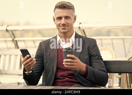 Il caffè mi completa. Il boss tiene la tazza da asporto e lo smartphone. Pausa caffè all'aperto. Bevanda calda. Tecnologia moderna. Colazione al mattino. Affari Foto Stock