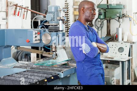 Uomo di lavoro afro-americano in tute blu accanto alla macchina per la lavorazione del vetro Foto Stock