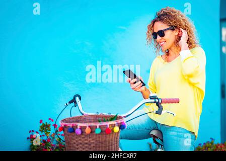 Ritratto di attraente adulto di mezza età femmina sorridente al telefono cellulare seduto su una bicicletta contro una parete blu sullo sfondo. La gente di donna gode technol Foto Stock