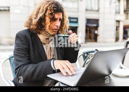 Uomo di mezza età con capelli ricci che bevono caffè caldo e navigano i dati sul laptop mentre lavora su progetto remoto in Street cafe Foto Stock