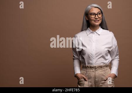 Vita in alto ritratto vista dell'elegante spensierata donna matura grigio-capelli posa alla fotocamera isolato su sfondo beige. Affascinante donna anziana sorridente toothy e guardando via Foto Stock