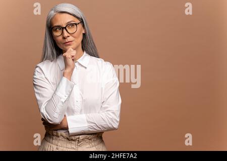 Donna matura intelligente e pensierosa che tiene il mento e ponderante idea, che prende la decisione difficile, che guarda incerta dubbia. Studio interno girato isolato su sfondo beige Foto Stock