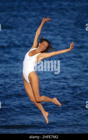 Abbastanza felice capelli marrone giovane donna che salta sulla spiaggia di sabbia bianco corpo vestito spruzzi onde blu cielo mare sfondo Foto Stock