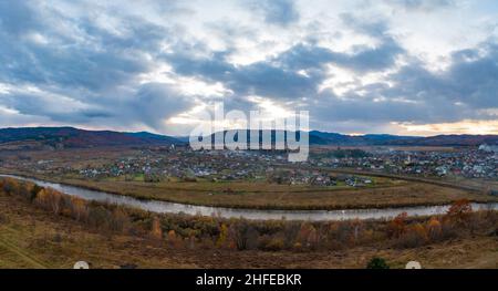 Stretto fiume che passa oltre l'insediamento tra la campagna autunnale contro alte montagne lontane sotto il cielo cupo con le nuvole piovose Foto Stock