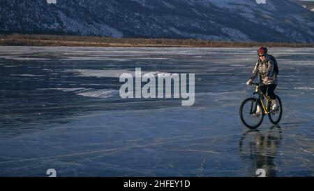 La donna sta pedalando in bicicletta sul ghiaccio. La ragazza è vestita con una giacca argentata in giù, zaino da bicicletta e casco. Ghiaccio del lago ghiacciato Baikal. Gli pneumatici in bicicletta sono coperti da punte. Il viaggiatore è in bicicletta. Foto Stock