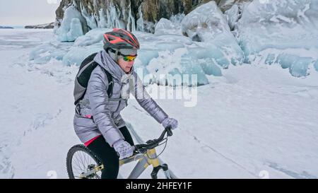 La donna va in bicicletta vicino alla grotta di ghiaccio. La roccia con grotte di ghiaccio e ghiaccioli. La ragazza è vestita con giacca argentata in giù, zaino da bicicletta e casco. Pneumatici invernali con punte. Il viaggiatore è in bicicletta. Foto Stock
