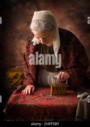 Vecchia donna in abito vittoriano che lavora con un ferro di carbone antico su un asse Foto Stock