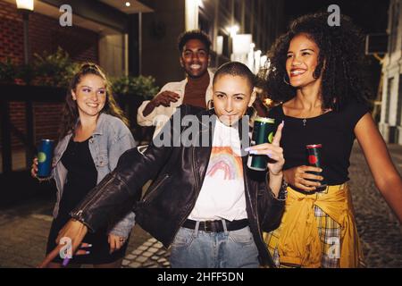 Gruppo di amici felici che ballano in città di notte. Quattro giovani vibranti che tengono lattine di birra mentre camminano insieme nella strada. Amici che hanno Foto Stock