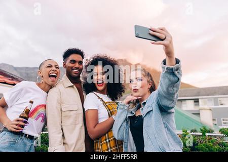 Amici che prendono un selfie di gruppo all'aperto. Quattro amici felici in posa per un selfie mentre si festeggiano insieme sul tetto. Gruppo di amici multiculturali Foto Stock