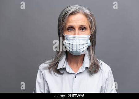 Donna anziana seria in abbigliamento formale e maschera medica sul viso isolato su grigio. Ritratto di primo piano di una dipendente di ufficio femminile che si protegge da malattie virali Foto Stock