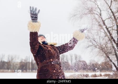 Donna anziana felice gettando neve nel nevoso parco invernale. Signora anziana che si diverte all'aperto durante la passeggiata indossando abiti caldi. Attività stagionali Foto Stock