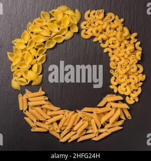 varietà di pasta cruda disposte in cerchio, isolate su sfondo di ardesia nera, dieta e concetto di cibo, vista dall'alto in basso con spazio copia Foto Stock