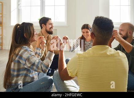 Gruppo di amici felici che si siedono in cerchio sul pavimento, tenendo le mani e ridendo Foto Stock