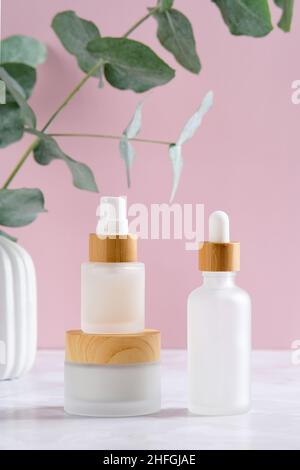 Eleganti contenitori cosmetici e eucalipto in vaso su sfondo rosa. Bottiglie d'olio essenziali e vasetto crema. Skincare prodotti di bellezza packaging design Foto Stock