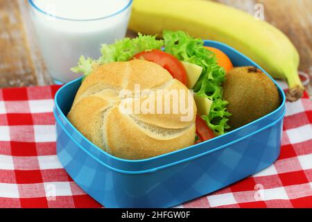 Un sano pranzo scolastico con rotolo di formaggio, lattuga e pomodoro, banana, kiwi e bicchiere di latte Foto Stock