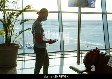 Uomo con power bank in aeroporto, ricarica il telefono per rimanere collegato durante il viaggio, prima del volo all'area di partenza. Passeggero con maschera facciale Foto Stock