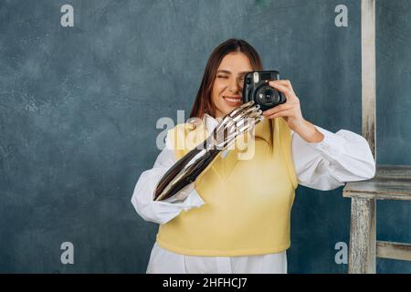 La giovane donna concentrata in camicia bianca e gilet con braccio bionico in metallo scatta foto con la moderna fotocamera su sfondo grigio Foto Stock