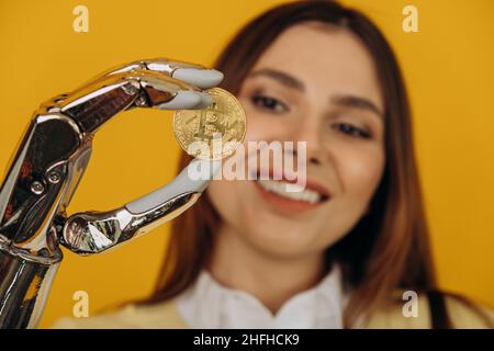 Lady con moderno braccio bionico in metallo lucido tiene grandi bitcoin dorati in posa per la fotocamera su sfondo giallo estrema vista ravvicinata Foto Stock
