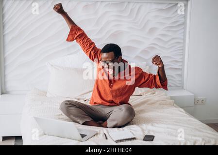 Il gioioso ragazzo afroamericano in abiti casual e occhiali mette le mani seduti vicino a un computer portatile aperto su un comodo letto a casa Foto Stock