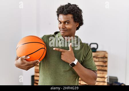 L'uomo africano con capelli ricci che tiene la palla di basket alla palestra sorridendo felice indicando con mano e dito Foto Stock