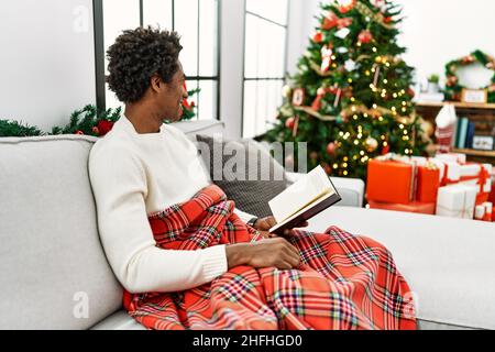 Giovane uomo africano americano che legge libro seduto sul divano da albero di natale a casa. Foto Stock