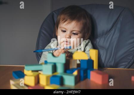 Ragazza del bambino che tiene una penna nelle sue mani mentre gioca con i blocchi della costruzione seduti in un high chair. Foto Stock