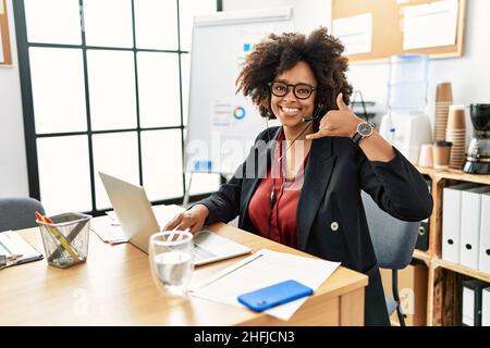 Donna afroamericana con capelli afroamericani che lavorano in ufficio indossando cuffie operatore sorridendo facendo il gesto del telefono con la mano e le dita come parlare Foto Stock