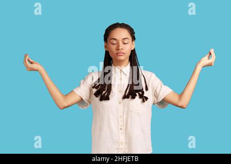 Donna calma e rilassata, con dreadlock neri in piedi con occhi chiusi, medita e gode di un'atmosfera tranquilla, fa mudra gesto, indossando camicia bianca. Studio interno girato isolato su sfondo blu. Foto Stock