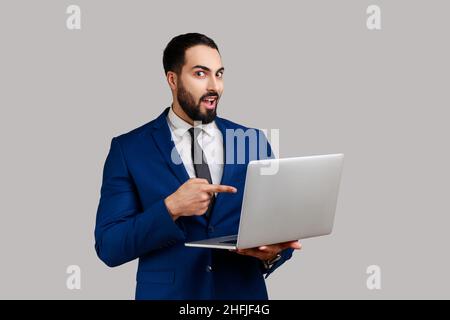 L'uomo sbalordito punta lo schermo del laptop e guarda la fotocamera con espressione scioccata, bocca aperta, vestendo il vestito di stile ufficiale. Studio interno girato isolato su sfondo grigio. Foto Stock