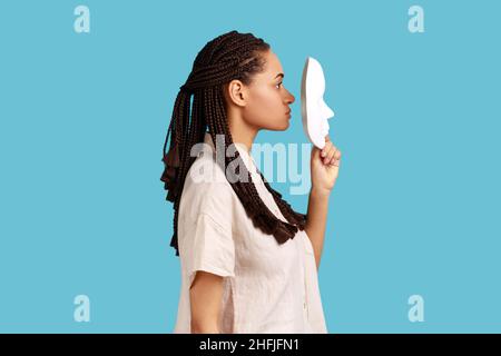 Ritratto della vista laterale della donna con i dreadlocks neri che tengono la maschera bianca davanti al suo viso, avendo espressione facciale rigorosa, indossando la camicia bianca. Studio interno girato isolato su sfondo blu. Foto Stock