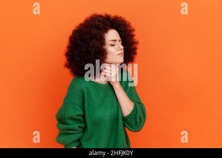 Ritratto del profilo di giovane donna adulta con acconciatura Afro indossando il maglione verde casual stile che ha mal di gola, toccando il collo, si sente male. Studio interno girato isolato su sfondo arancione. Foto Stock