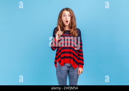 Giovane donna emozionata che indossa un pullover casual a righe in piedi con un dito sollevato, con un'idea improvvisa, guardando la fotocamera a bocca aperta. Studio interno girato isolato su sfondo blu. Foto Stock