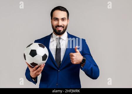 Uomo d'affari soddisfatto bearded che tiene la sfera di calcio sulla sua mano con espressione positiva sorridente, mostrando il pollice in su, indossando il vestito ufficiale di stile. Studio interno girato isolato su sfondo grigio. Foto Stock