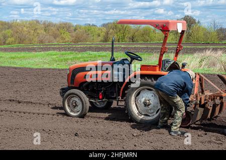 Lavoro agricolo di primavera nel campo, piantando patate nel villaggio. Trattore piccolo rosso con un aratro e un uomo dal retro. Foto Stock