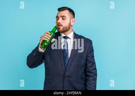 Uomo d'affari giovane adulto portatore di vestito di stile ufficiale che beve alcool, che tiene la bottiglia di birra in mano, che soffre di alcolismo. Studio interno girato isolato su sfondo blu. Foto Stock