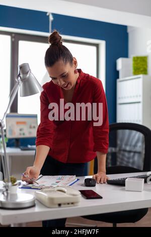 Donna d'affari in piedi vicino alla scrivania in ufficio startup guardando i grafici. Imprenditore che si appoggia sopra gli appunti con i documenti di affari. Dipendente in camicia rossa che guarda i grafici dei dati sul tavolo. Foto Stock