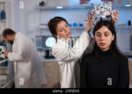 Neurologo medico donna mettere eeg scanner sulla testa del paziente analisi dell'evoluzione del cervello lavorando alla diagnosi di malattia durante l'esperimento neurologico in laboratorio high tech. Dottore scienziato che spiega la tomografia Foto Stock