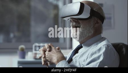 Un uomo d'affari maturo che indossa un visore VR e sperimenta la realtà virtuale nel suo ufficio Foto Stock
